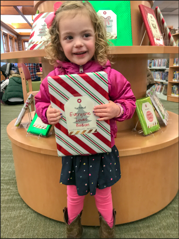 A little girl is excited to unwrap a book wrapped in shiny white, red and green paper with a tag that reads 
