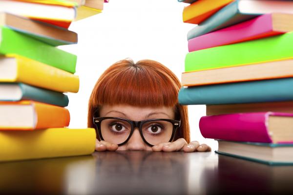 a girl teen with red hair, bangs, and glasses peeking through a stack of books